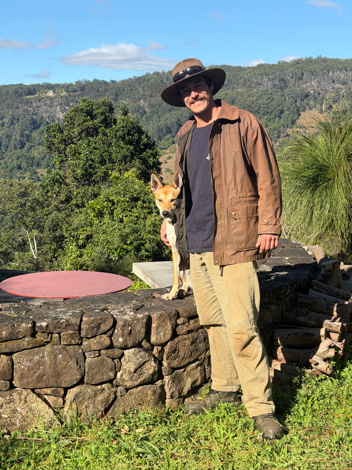 Calvin Pullen standing with Wispah the dingo in a garden with forested hills behind them.